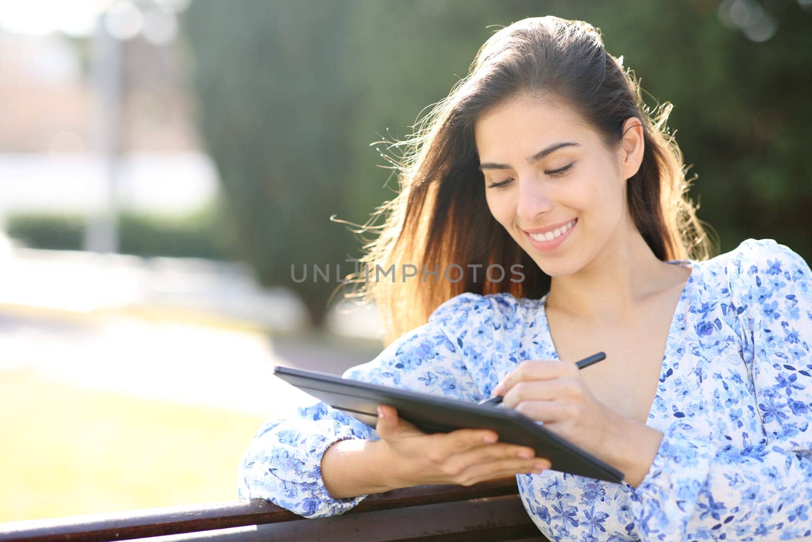 Happy woman writing on tablet with pen sitting in a park