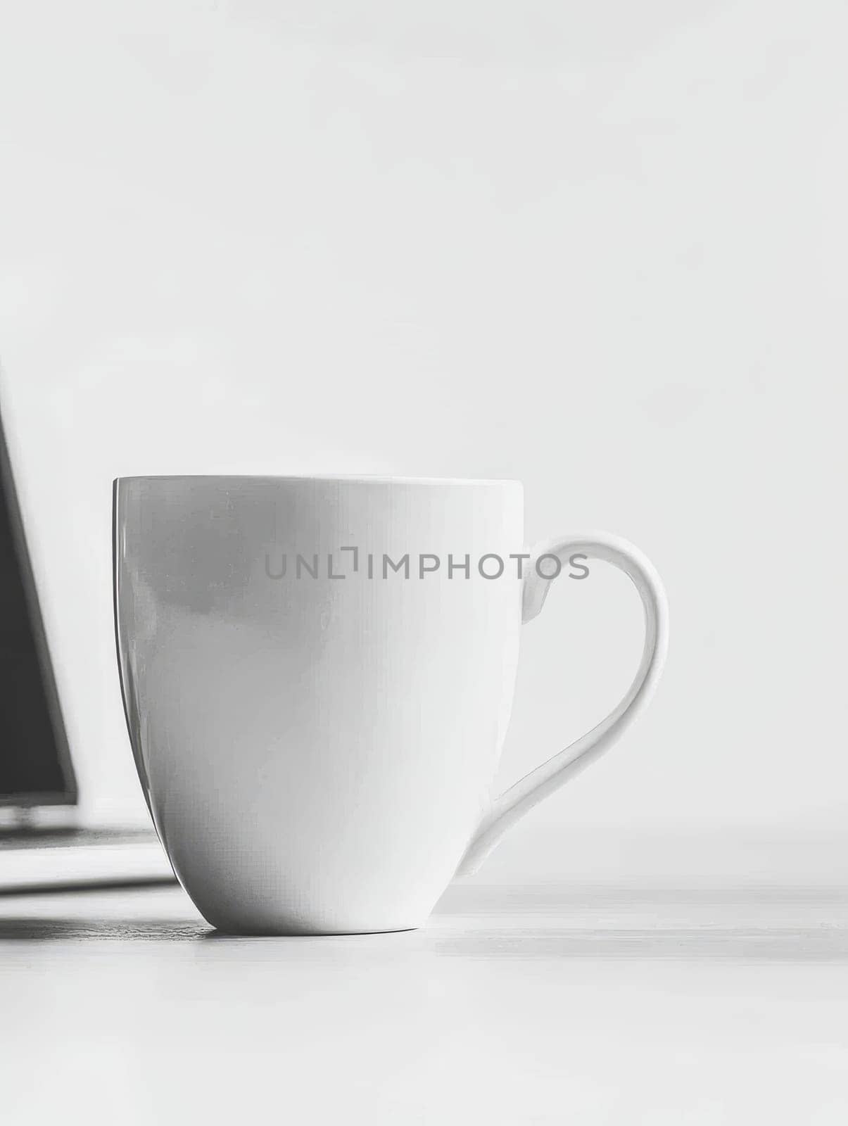 A simple white ceramic coffee mug sits on a light surface next to a blurred laptop, embodying a clean and modern workspace aesthetic.