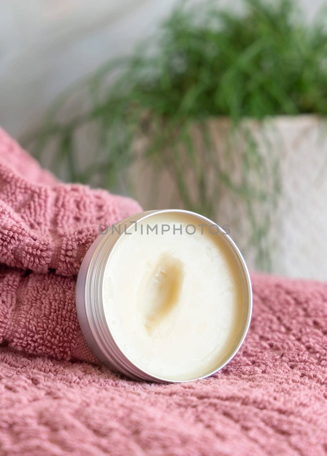 Opened body butter jar on pink towel against green plant in bathroom, close up. Natural herbal cosmetic product for body care, everyday lip care routine