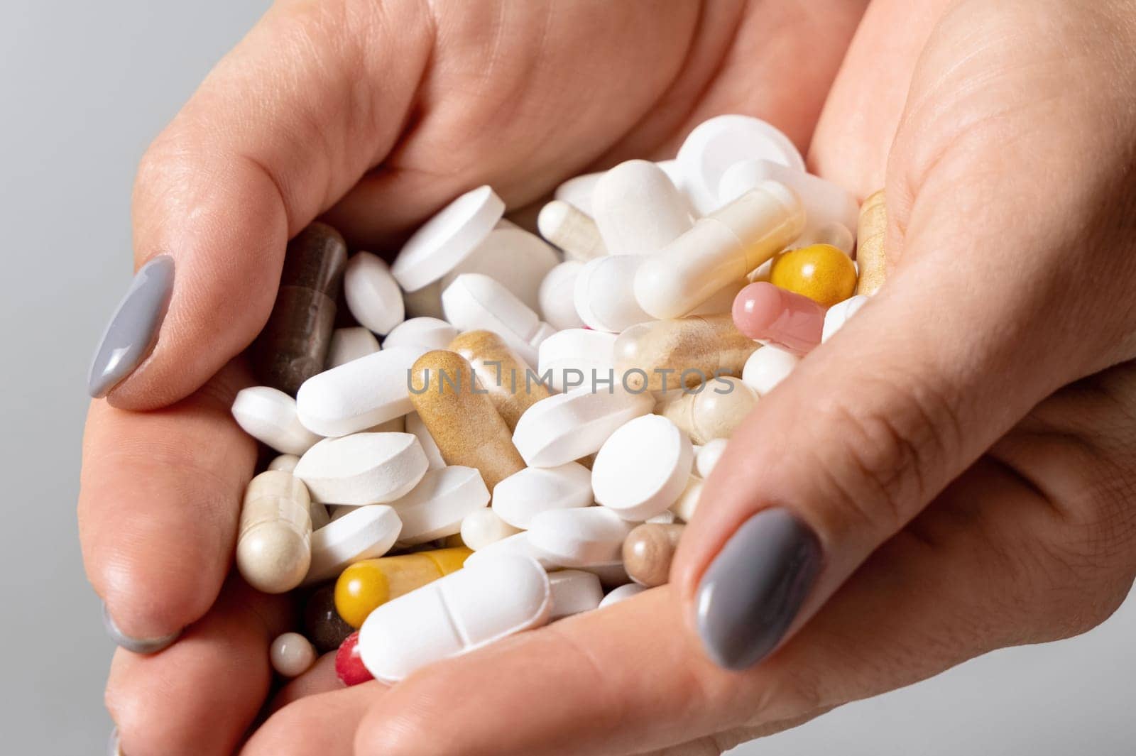 Cupped womans hands with a pile of mixed white and beige medical pills, tablets and capsules closeup over grey background. Assorted pharmaceutical products for health