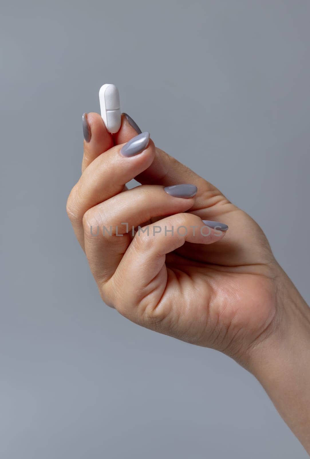 Female hand holding a white pill with fingers closeup against grey background. Healthcare, medical and pharmaceutical concept.