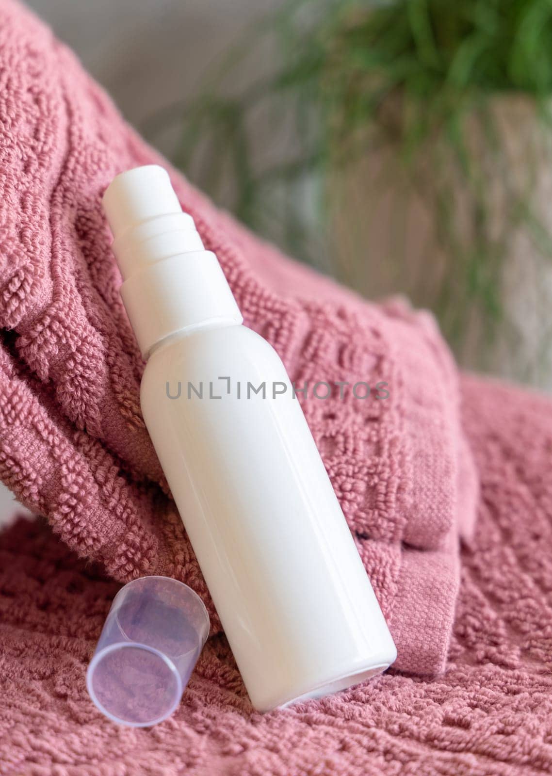 White cosmetic spray bottle on pink folded towels near green plant in bathroom, close up, packaging mockup. Natural face cream for personal care, toner, tonic or micellar water