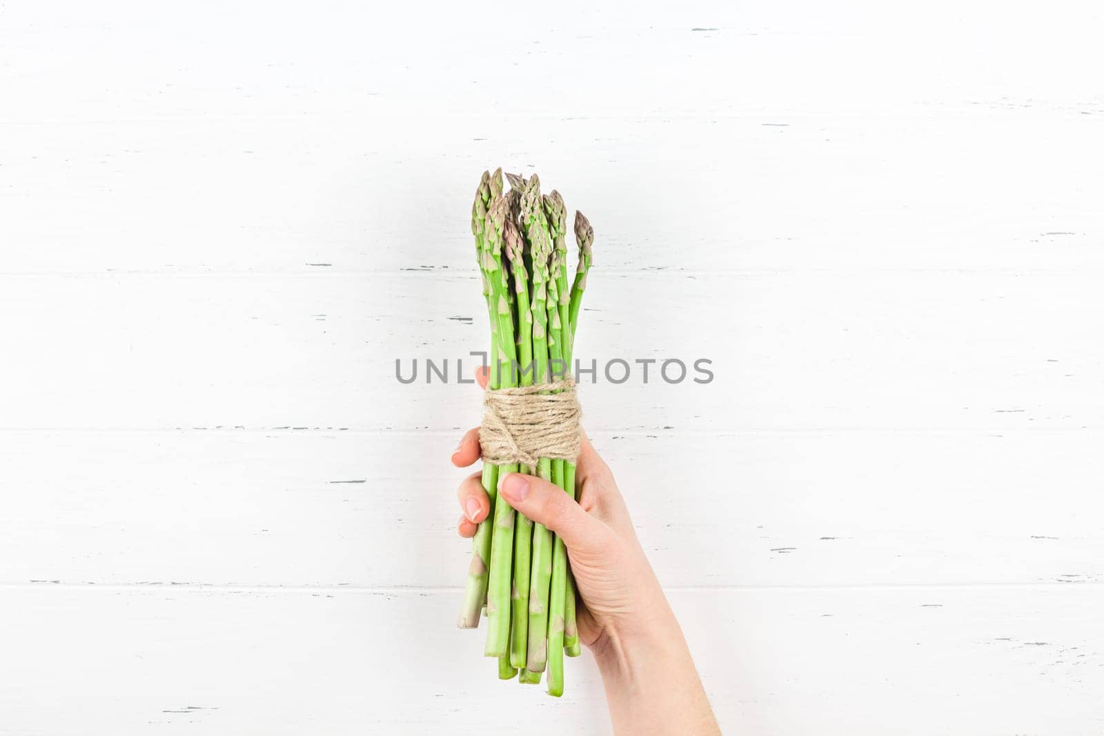 Creative scandinavian style flat lay top view of fresh green asparagus in woman hands on white wooden table background copy space. Minimal house cooking concept for blog or recipe book