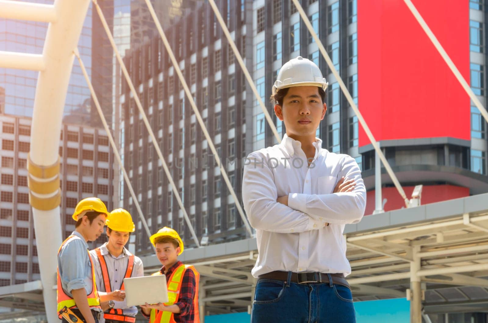 Portrait of Asian male project engineer in white helmet standing with crossed arms in front of modern building with three men working with laptop on background