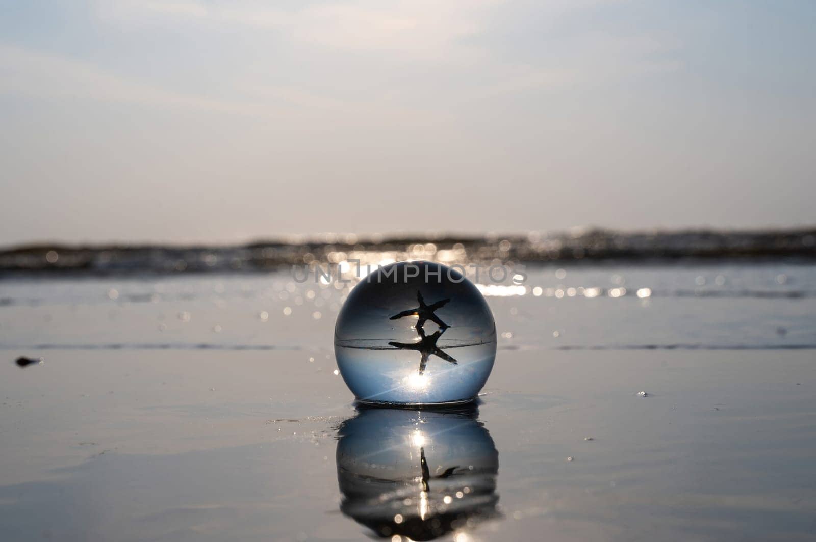 A glass ball lies in the waves on the sandy beach, the ball reflects the sea, a starfish and the setting sun