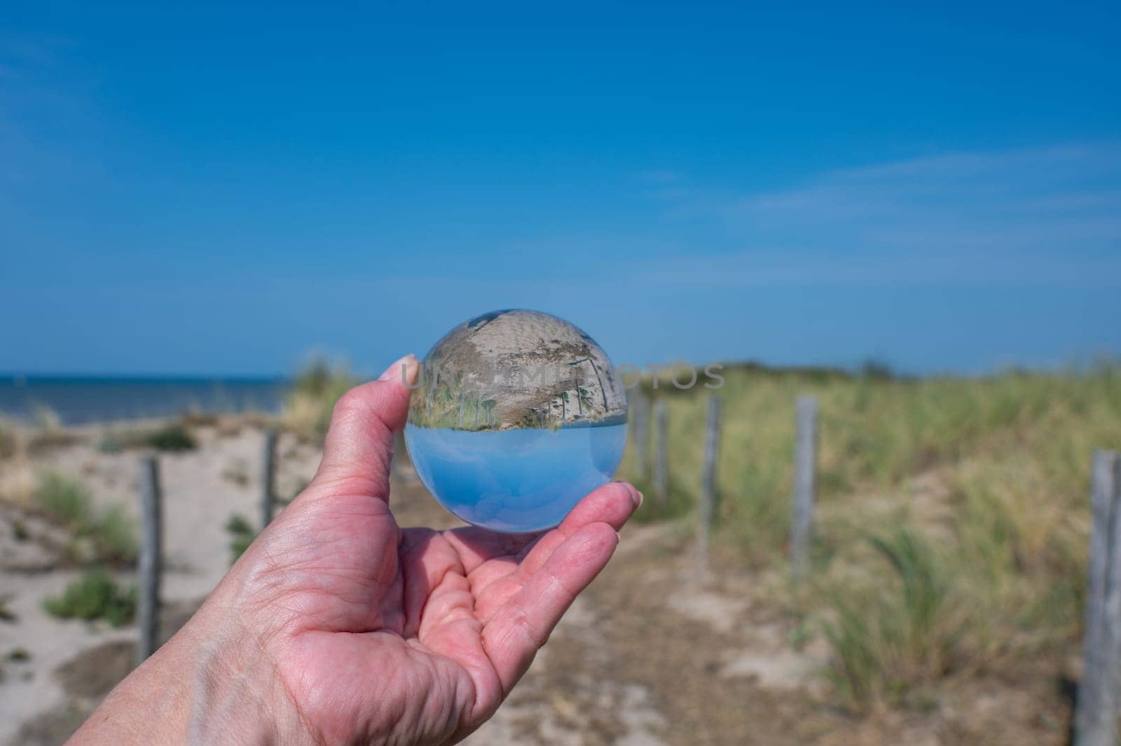 A human hand holds a glass ball, in the ball a sand dune, a footpath, blue sky and the sea are reflected