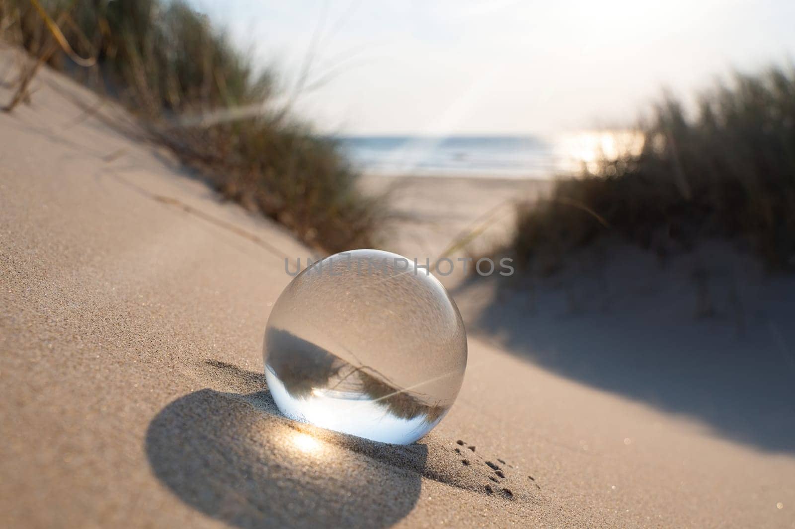 A glass ball lies in the sand , the sea and dune grass  are reflected in the ball