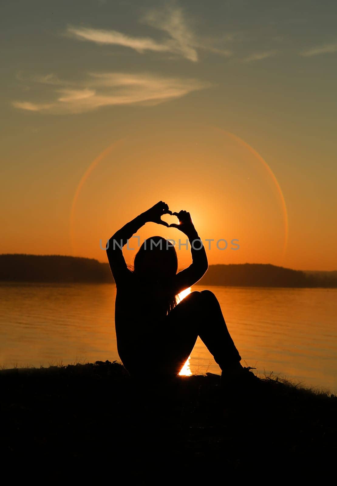 The silhouette of a girl holds her fingers in the shape of a heart against the background of a bright sunset over shimmering water
