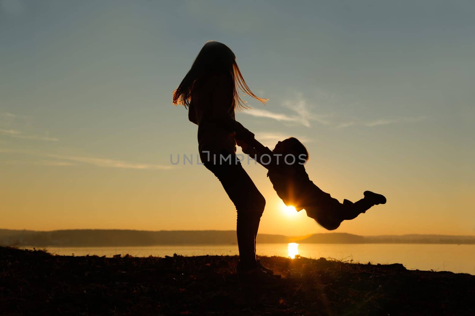 The silhouette perfectly captures the image of a girl lifting her brother in her arms against the background of a sunset by the water