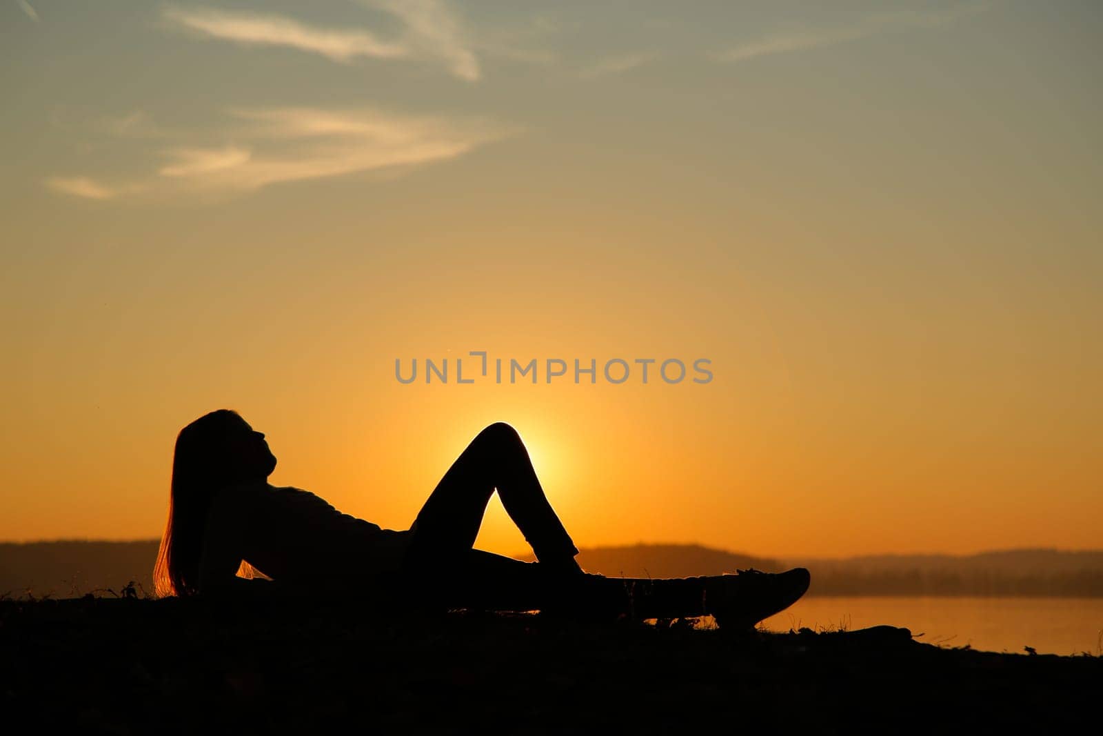 A beautifully serene silhouette of a woman peacefully lying by the calm water during a stunning sunset