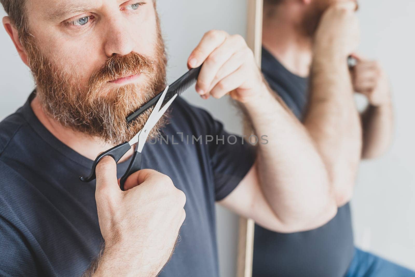 Selfcare during quarantine isolation. Adult handsome caucasian man cutting moustache and beard himself personally with scissors at home. Coronavirus outbreak and people shutdown, stay home concept