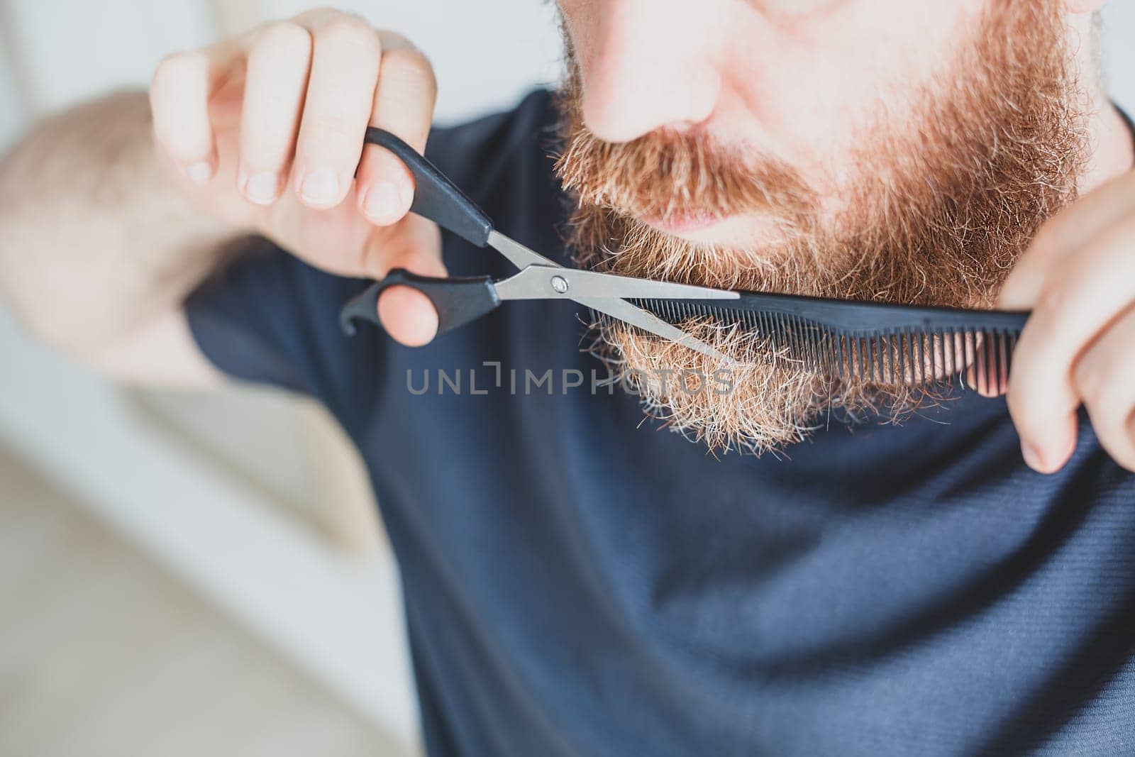 Selfcare during quarantine isolation. Adult handsome caucasian man cutting moustache and beard himself personally with scissors at home. Coronavirus outbreak and people shutdown, stay home concept