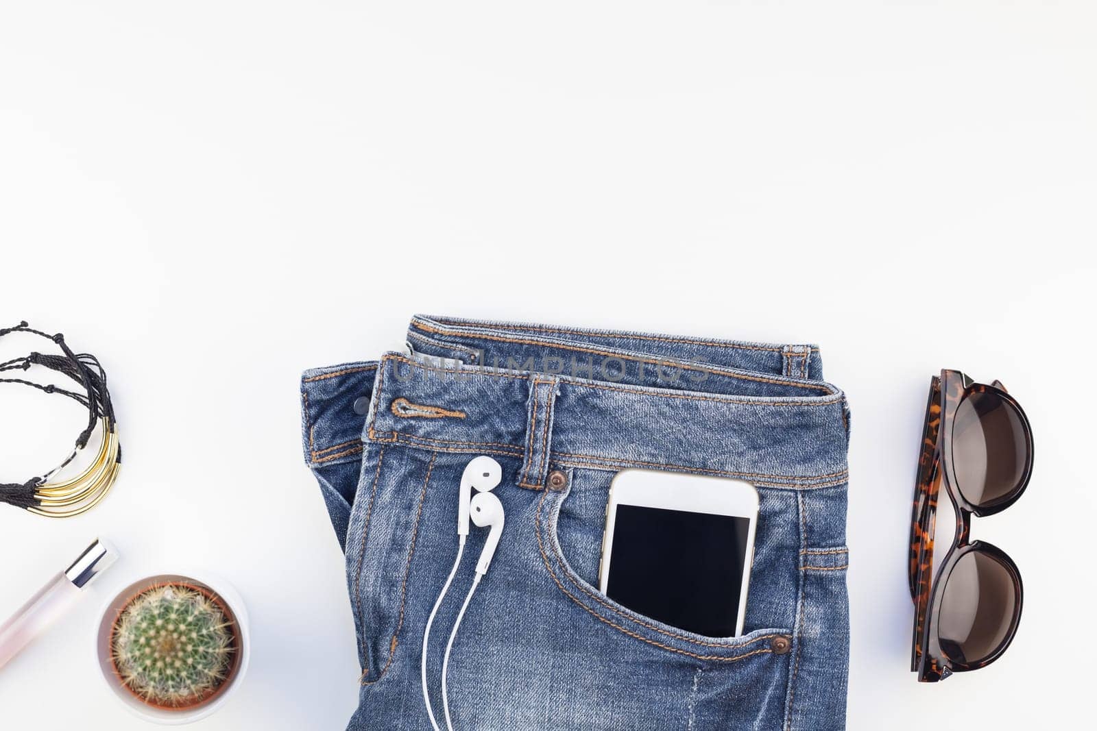 Flat lay of suede shoes, blue jeans, bracelets and smartphone with headphones on white background with copy space. Overhead view of woman casual outfit. Trendy hipster look top view