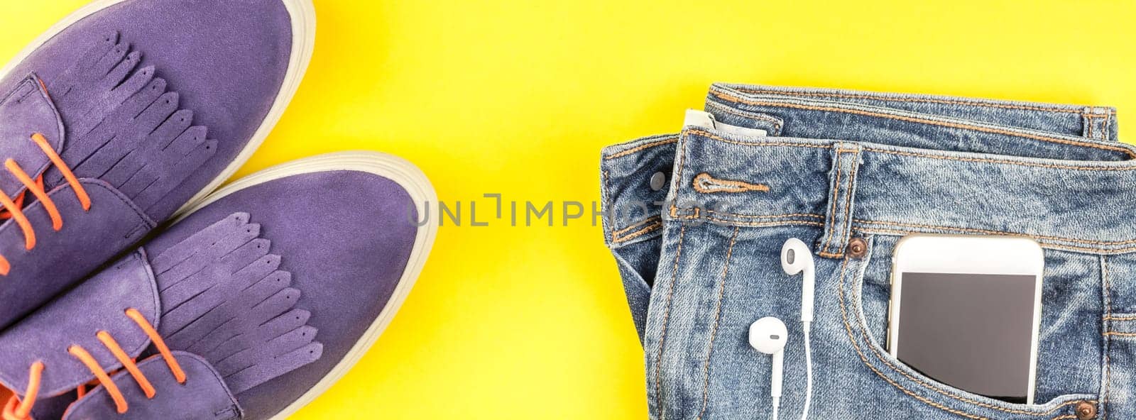 Flat lay of suede shoes, blue jeans, grey hat, cactus and smartphone headphones on bold yellow paper background with copy space. Overhead view of woman casual outfit. Long wide banner