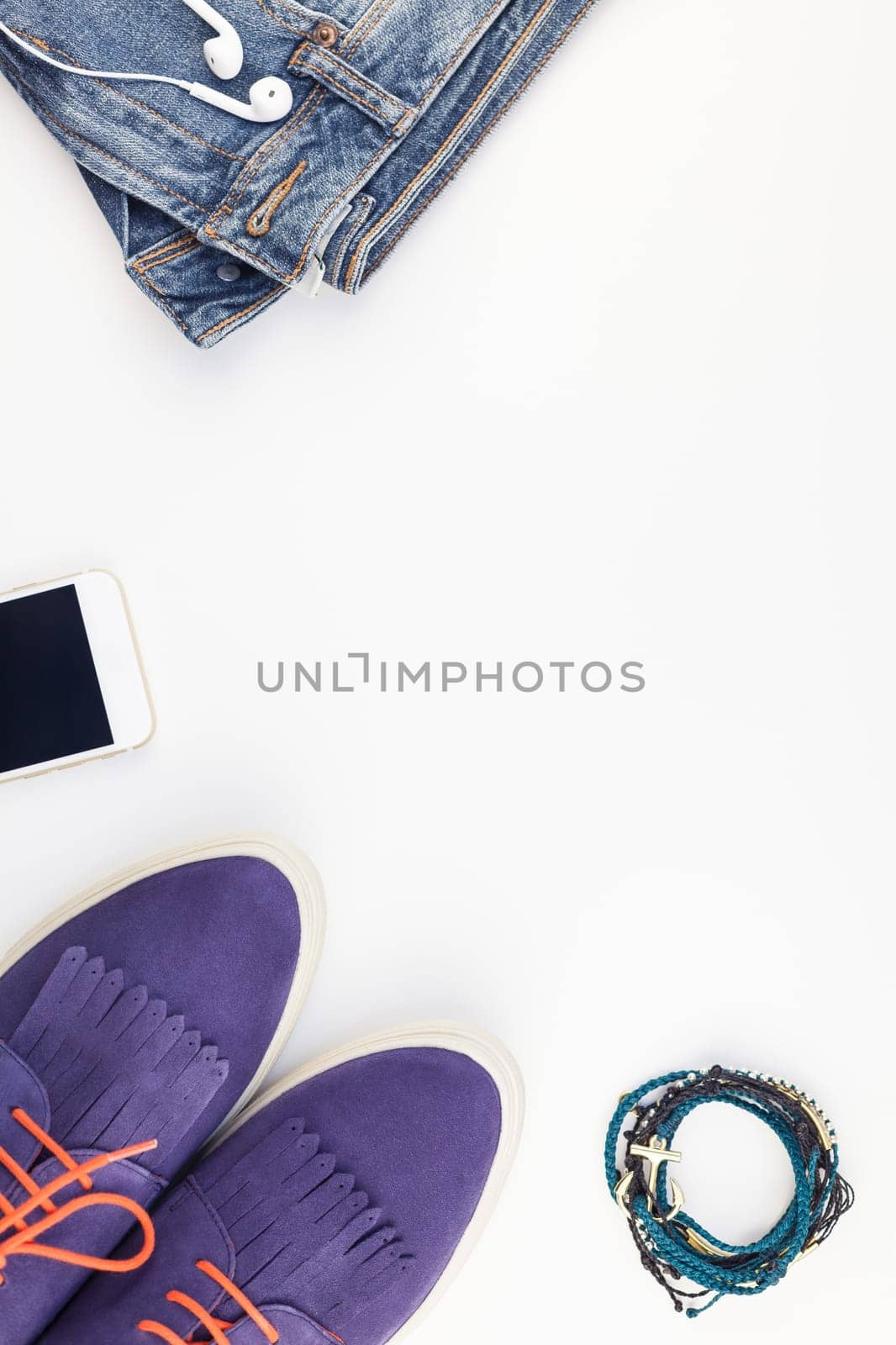 Flat lay of suede shoes, blue jeans, bracelets and smartphone with headphones on white background with copy space. Overhead view of woman casual outfit. Trendy hipster look top view