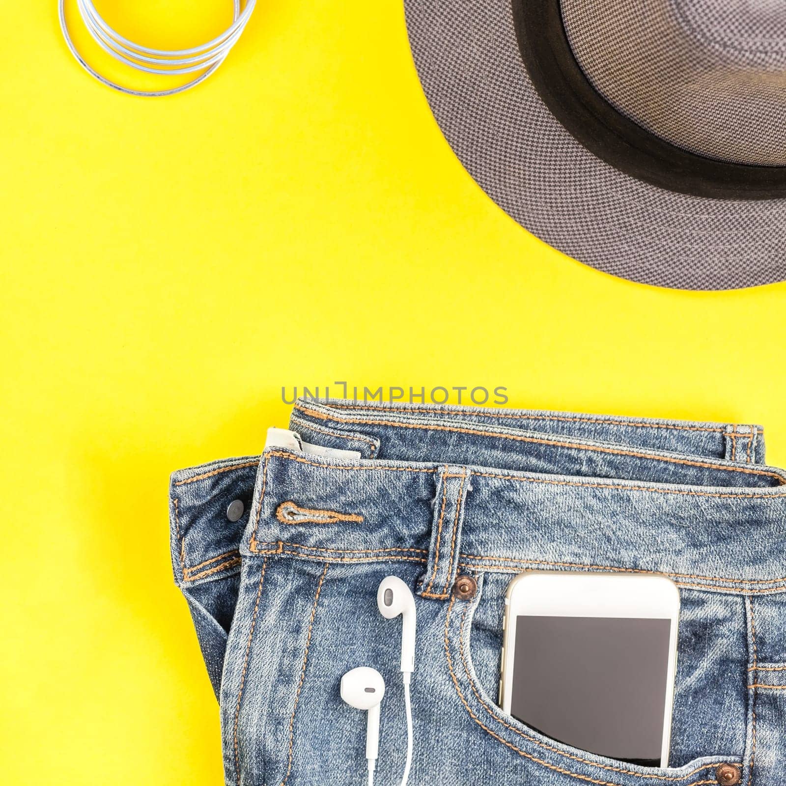 Flat lay of suede shoes, blue jeans, grey hat, cactus and smartphone headphones on bold yellow paper background with copy space. Overhead view of woman casual outfit. Square hipster look top view