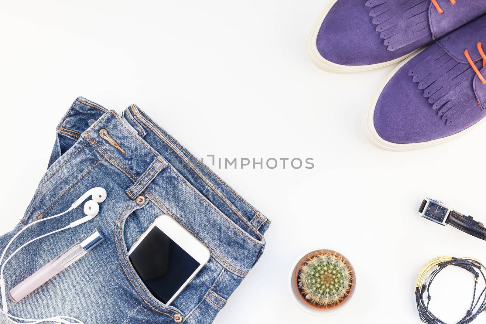 Flat lay of suede shoes, blue jeans, bracelets cactus and smartphone with headphones on white background with copy space. Overhead view of woman casual outfit. Trendy hipster look top view
