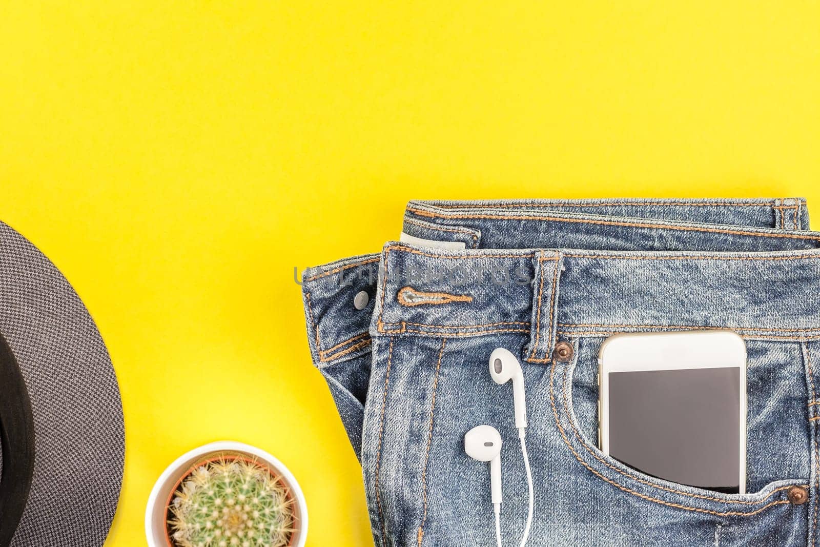 Flat lay of blue jeans, grey hat, cactus and smartphone headphones on bold yellow paper background with copy space. Overhead view of woman casual outfit. Trendy hipster look top view