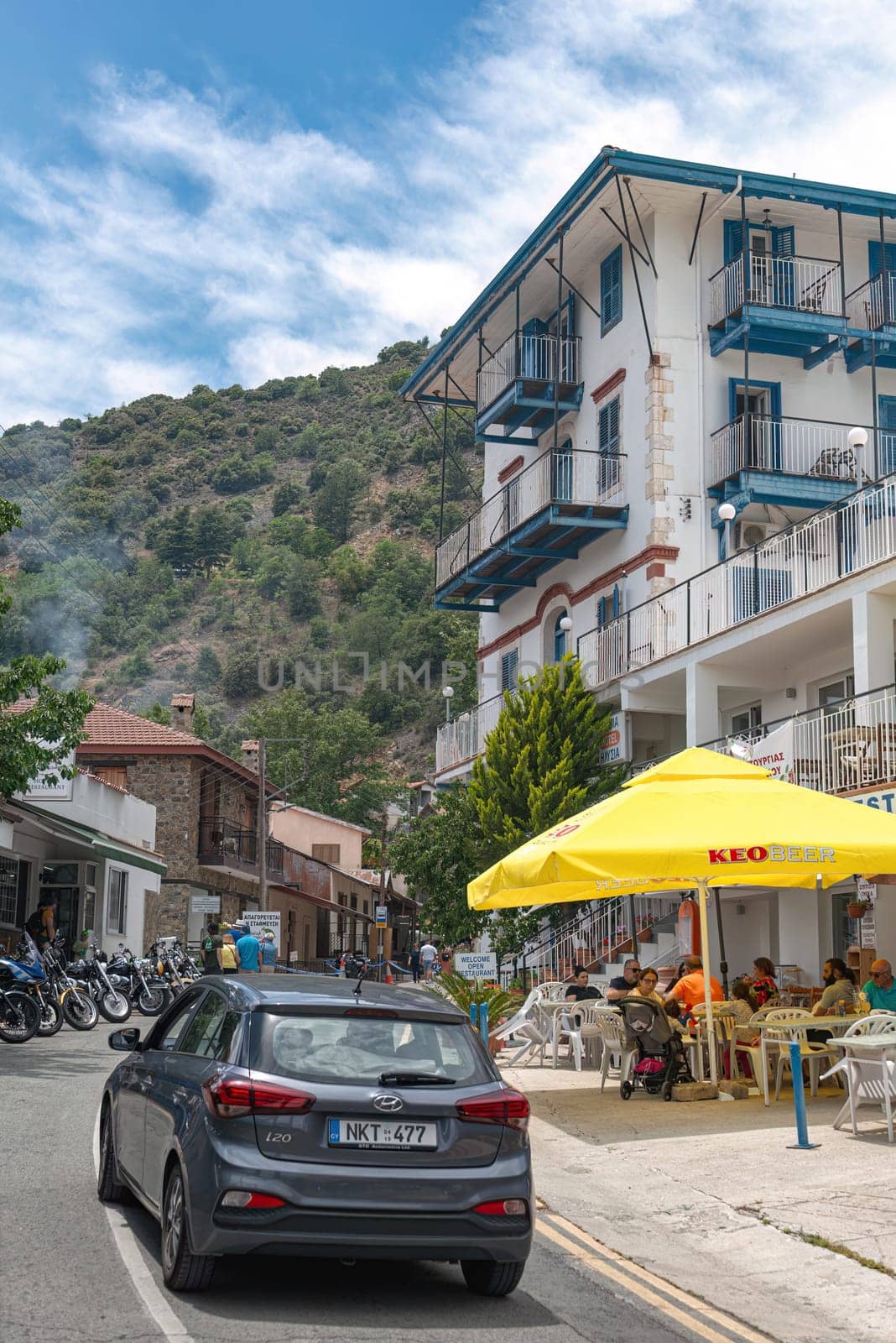 Pedoulas, Limassol District, Cyprus - June 11, 2023: Car driving on a narrow street in Pedoulas village, Cyprus, passing by tourists enjoying their meal