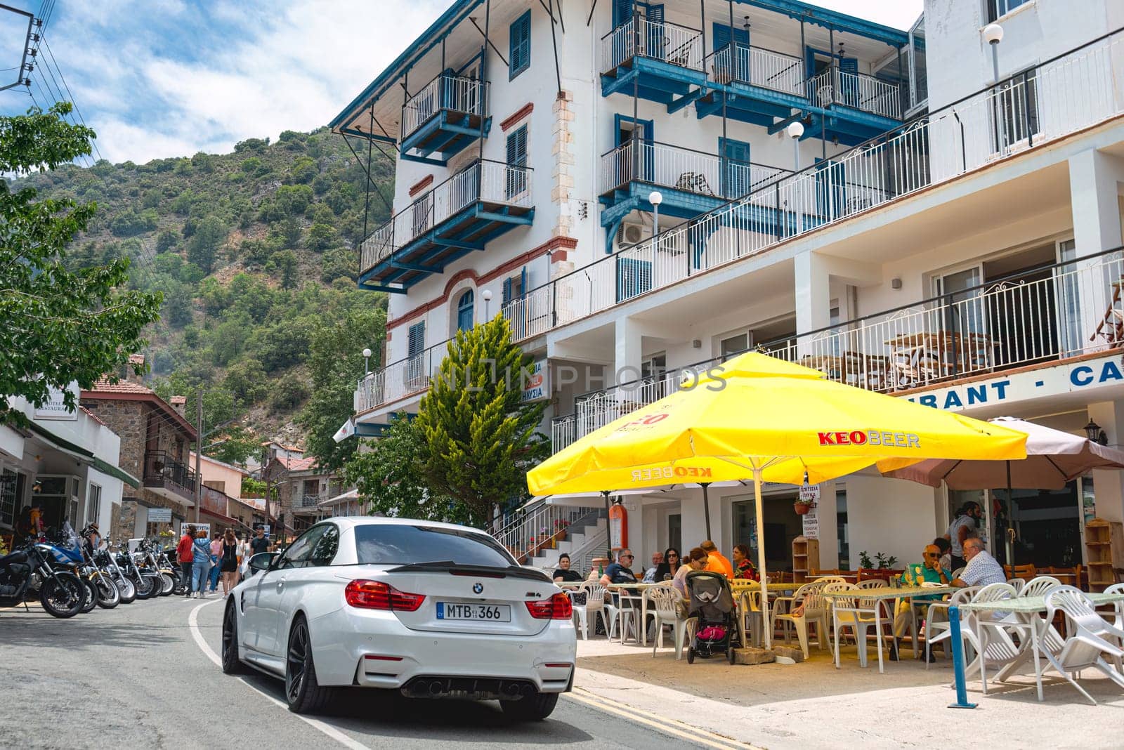 Pedoulas, Limassol District, Cyprus - June 11, 2023: White convertible driving on a narrow street, passing outdoor restaurant terraces with customers enjoying their meals in Pedoulas village, Cyprus
