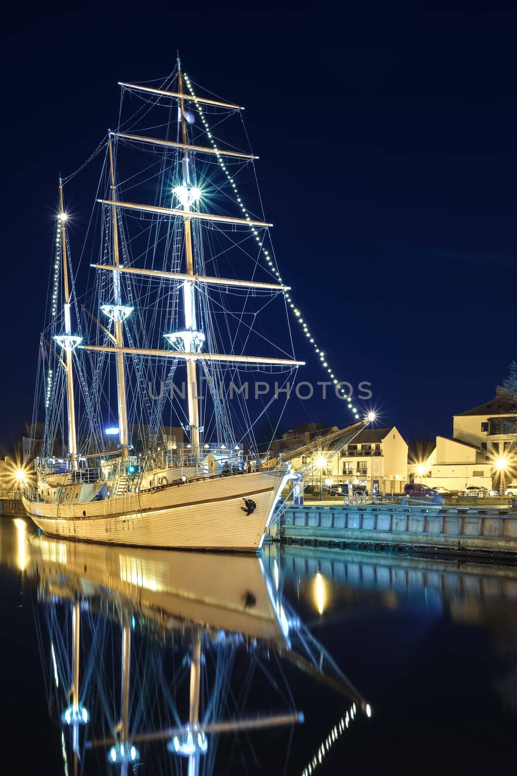 Sailing boat anchored in old town of Klaipeda, Lithuania, with evening lights on. Medieval town and barquentine reflection in calm waters.