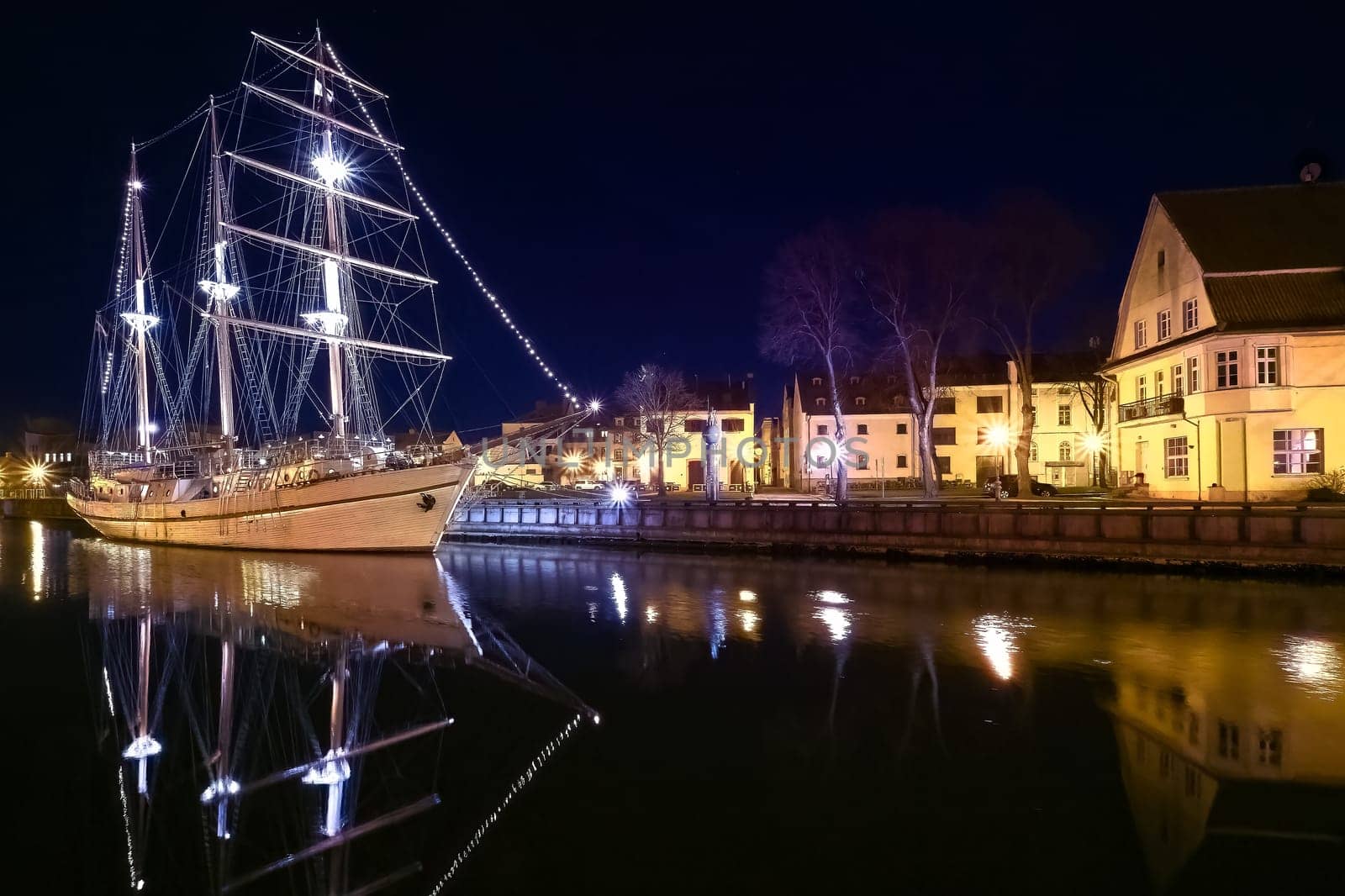 Sailing boat anchored in old town of Klaipeda, Lithuania, with evening lights on. Medieval town and barquentine reflection in calm waters.