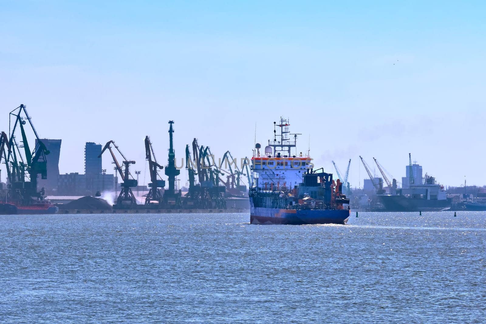 View of commercial cargo port and its container bridge cranes and a ship entering port on a sunny day.