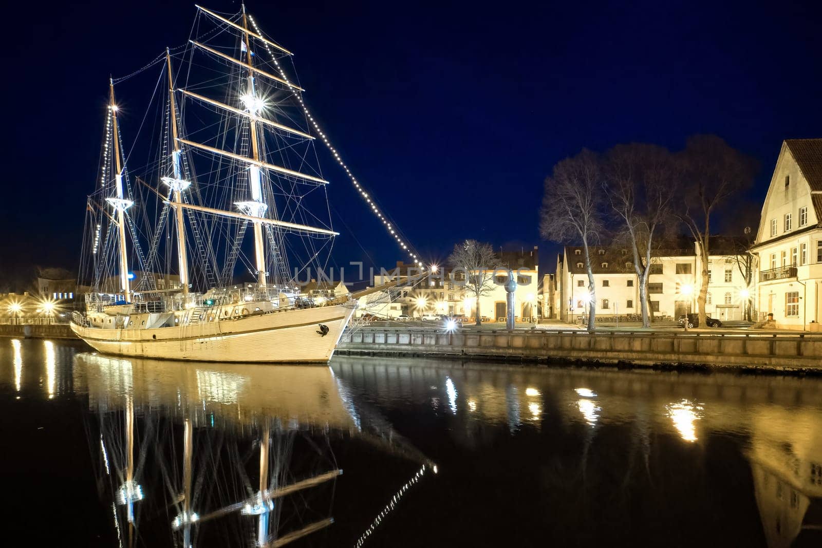 Sailing boat anchored in old town of Klaipeda, Lithuania, with evening lights on. Medieval town and barquentine reflection in calm waters.