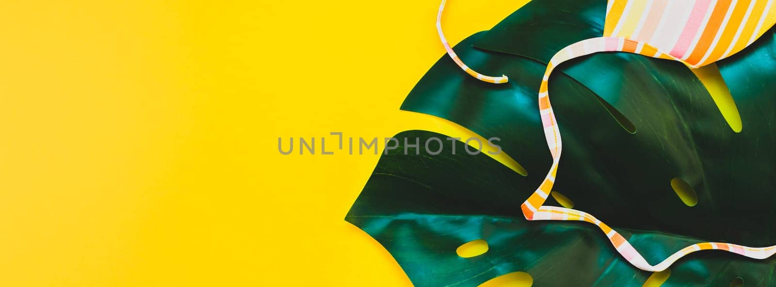Tropical jungle palm monstera leaves and a woman's swimsuit top on bright yellow paper background. Summer vacations creative flat lay concept template for text. Long wide banner