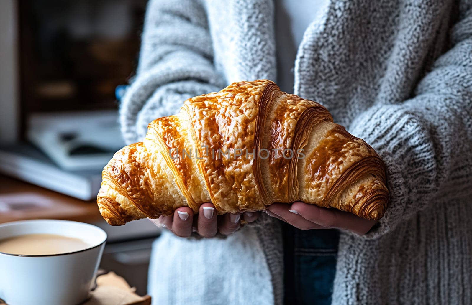 A person holds a freshly baked croissant with golden layers, while coffee awaits in a cozy kitchen.