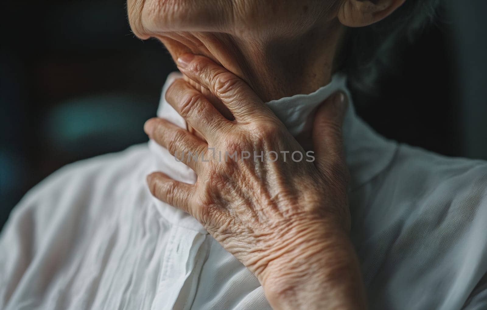 An older individual thoughtfully touches their throat, dressed in a white collared shirt, in a softly lit room.