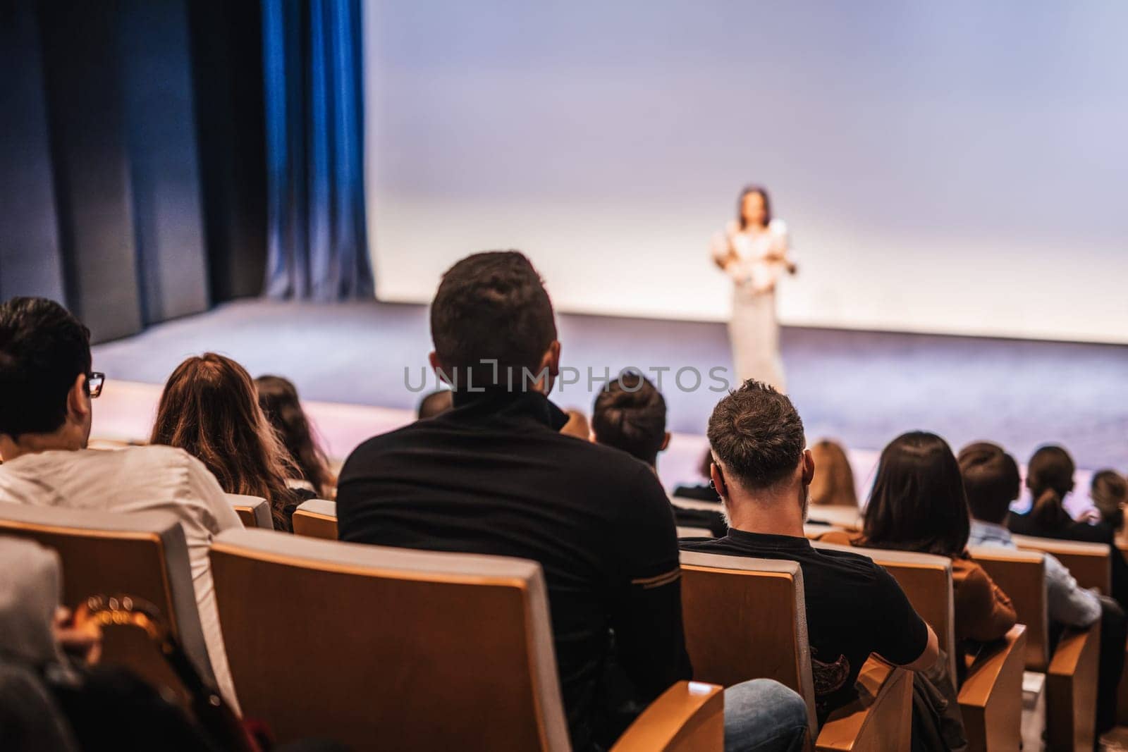Business and entrepreneurship symposium. Female speaker giving a talk at business meeting. Audience in conference hall. Rear view of unrecognized participant in audience.