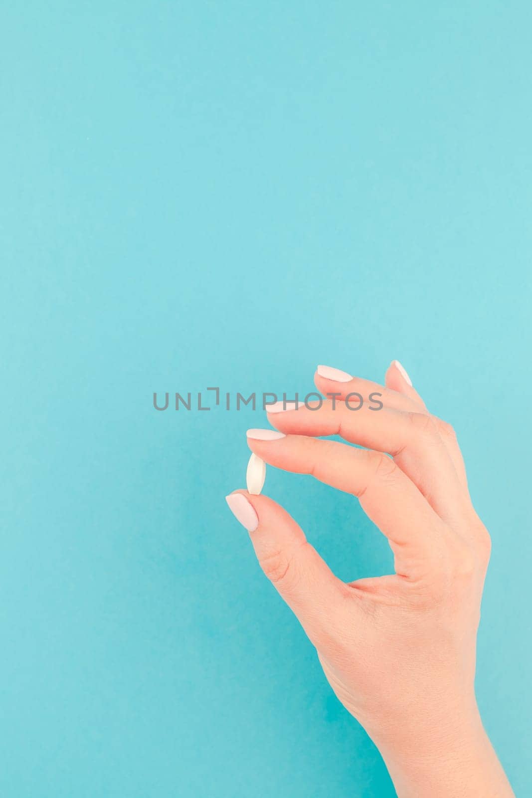 Woman hand holding a white pill on pastel blue paper background with copy space in minimal style, template for text. Concept of medical treatment, pharmacy, healthcare.