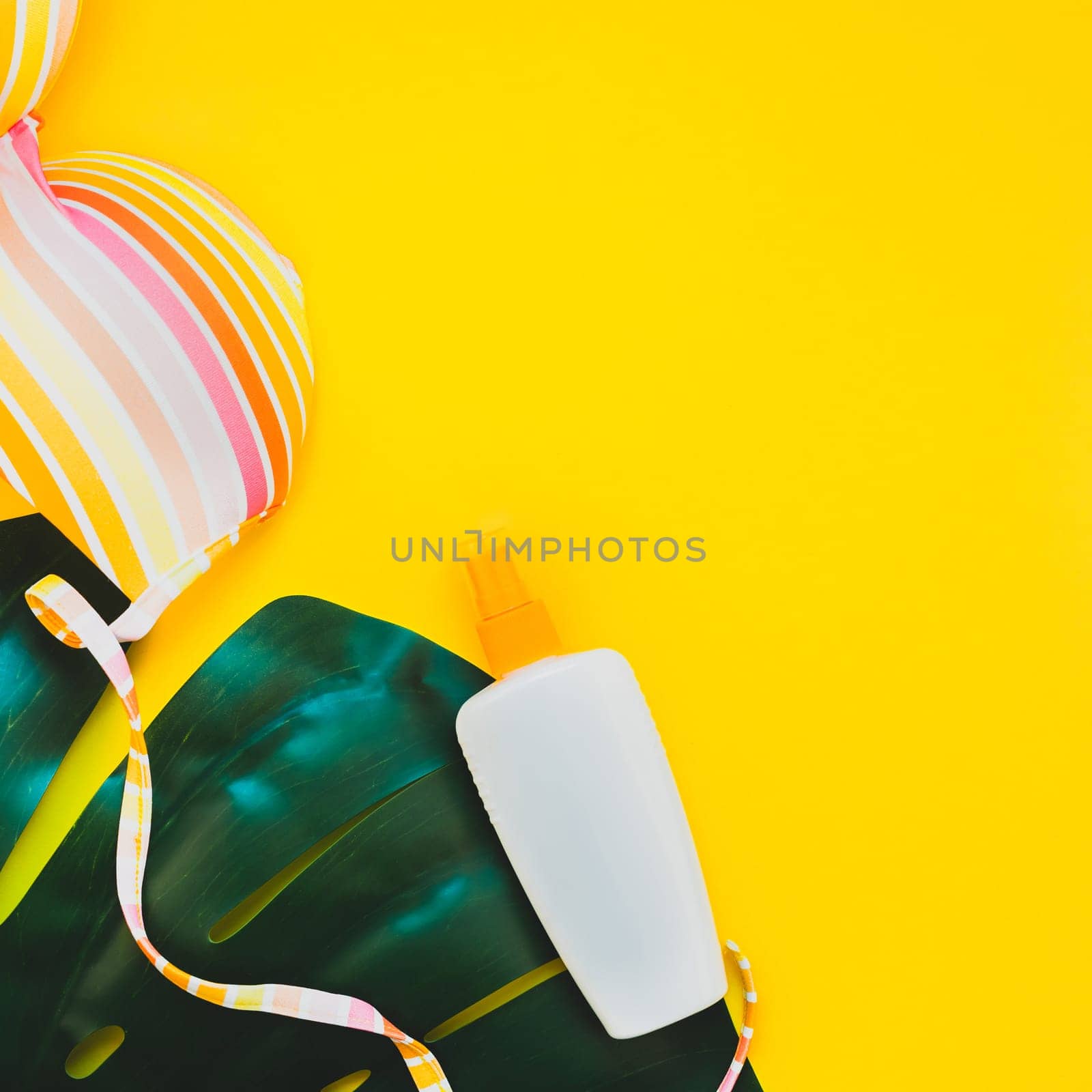 Tropical jungle palm monstera leaves, sunglasses, sunscreen blank bottle mockup and woman's swimsuit top on bright square yellow paper background. Summer vacations creative flat lay concept template