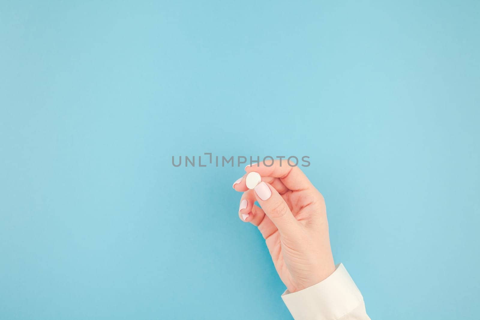 Woman hand holding a white pill on pastel blue paper background with copy space in minimal style, template for text. Concept of medical treatment, pharmacy, healthcare.