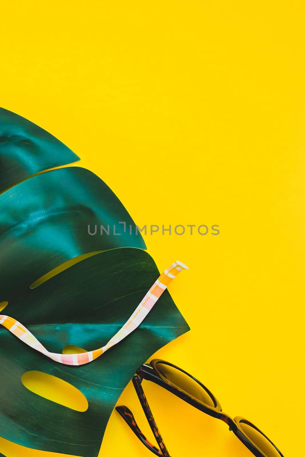 Tropical jungle palm monstera leaves, sunglasses and a woman's swimsuit top on bright yellow paper background. Summer vacations creative flat lay concept template for text