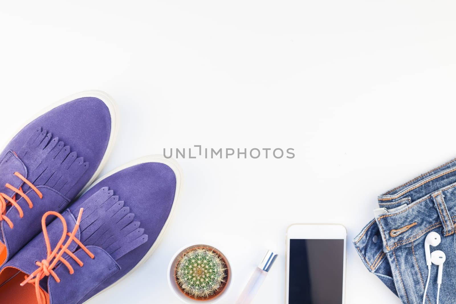 Flat lay of suede shoes, blue jeans, bracelets cactus and smartphone with headphones on white background with copy space. Overhead view of woman casual outfit. Trendy hipster look top view