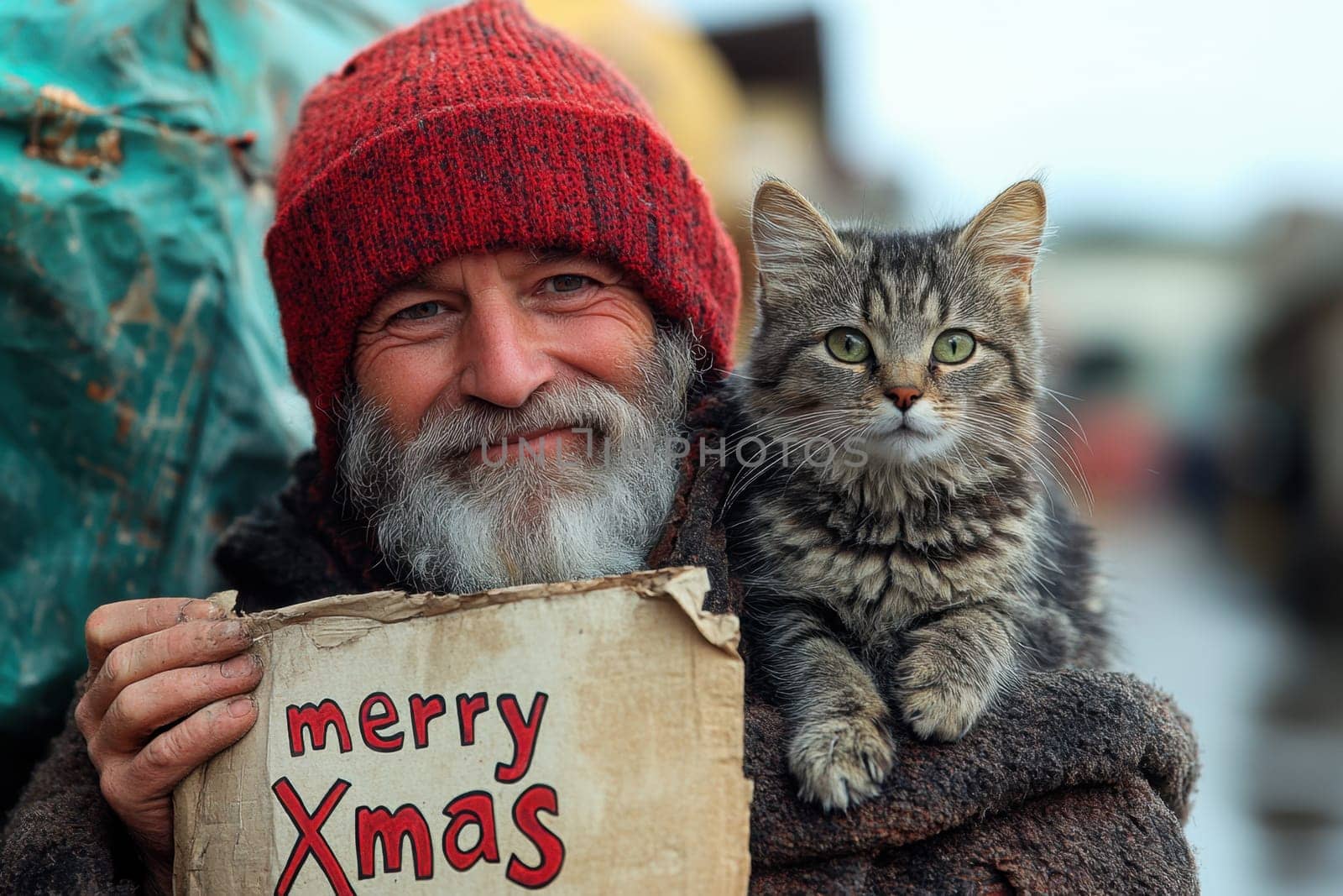A joyful man wearing a red hat and warm clothing stands outside with a cat on his shoulder holding a cheerful Christmas sign.