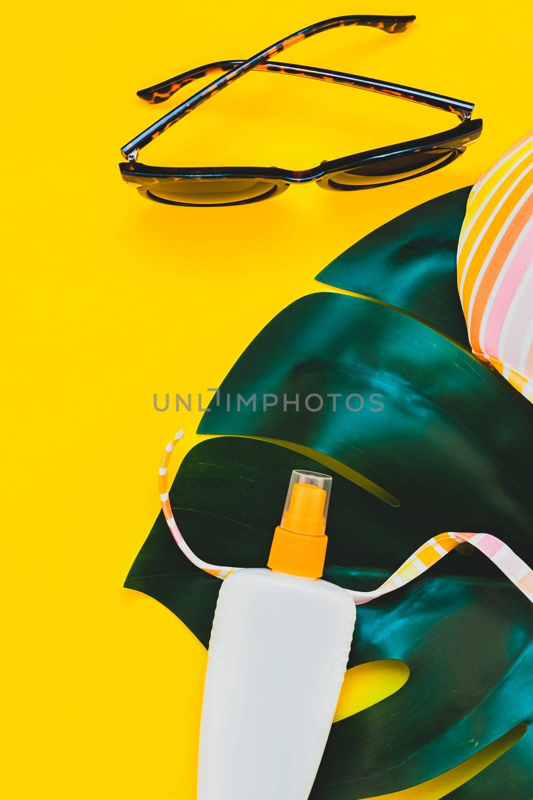 Tropical jungle palm monstera leaves, sunglasses, sunscreen blank bottle mockup and woman's swimsuit top on bright yellow paper background. Summer vacations creative flat lay concept template for text