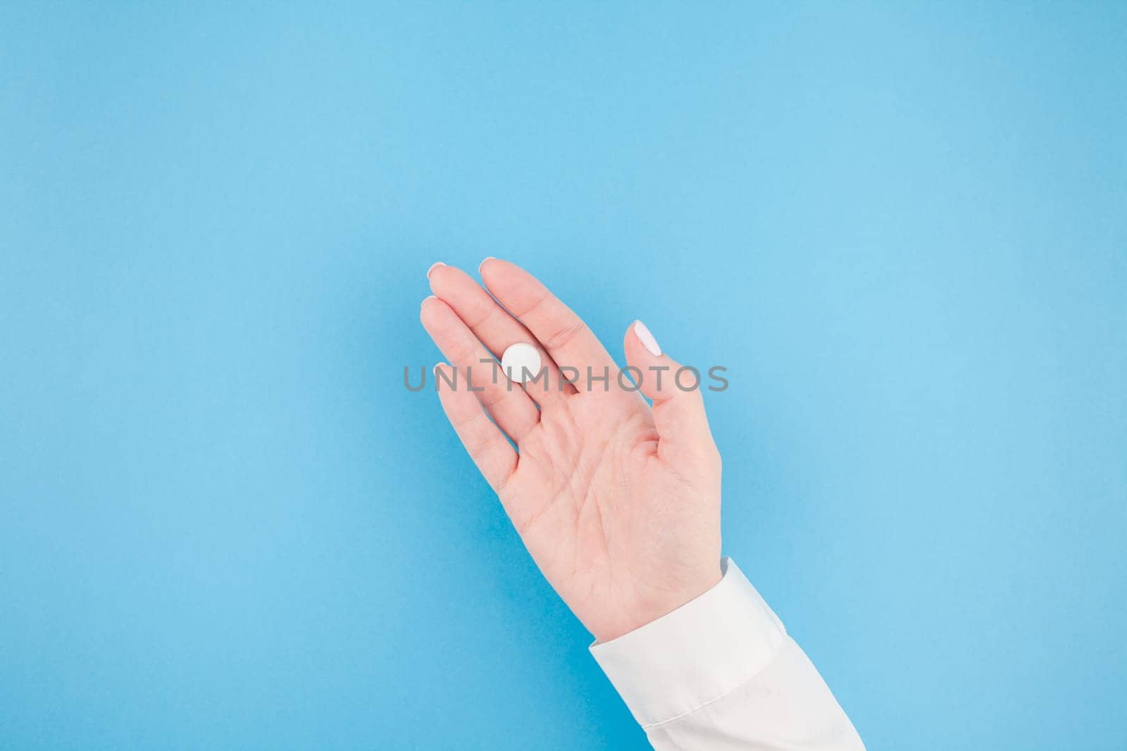 Woman hand holding a white pill on pastel blue paper background with copy space in minimal style, template for text. Concept of medical treatment, pharmacy, healthcare.