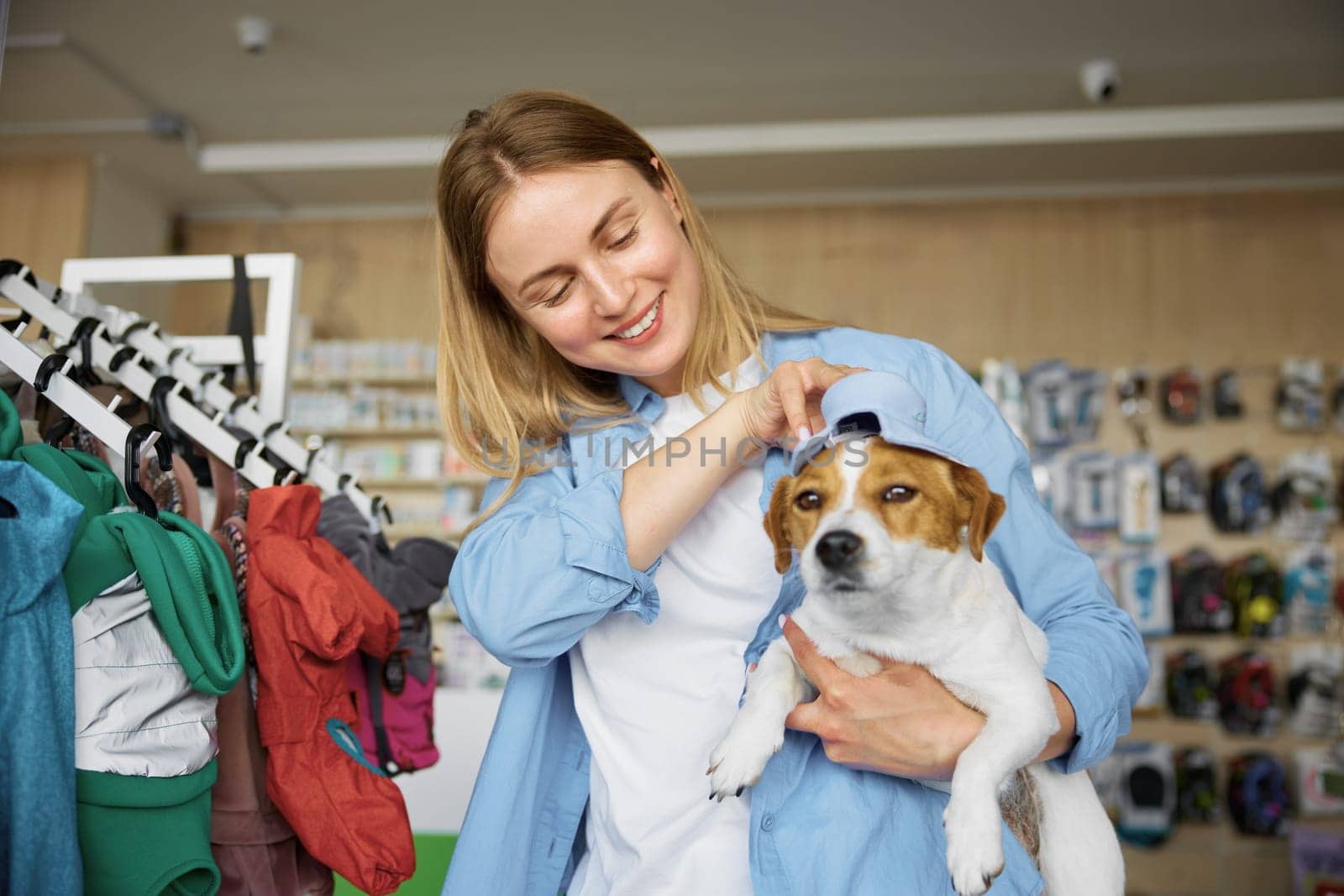 Young woman putting on cute costume hat on adorable dog head enjoying shopping at pet shop. Seasonal purchases for domestic animal