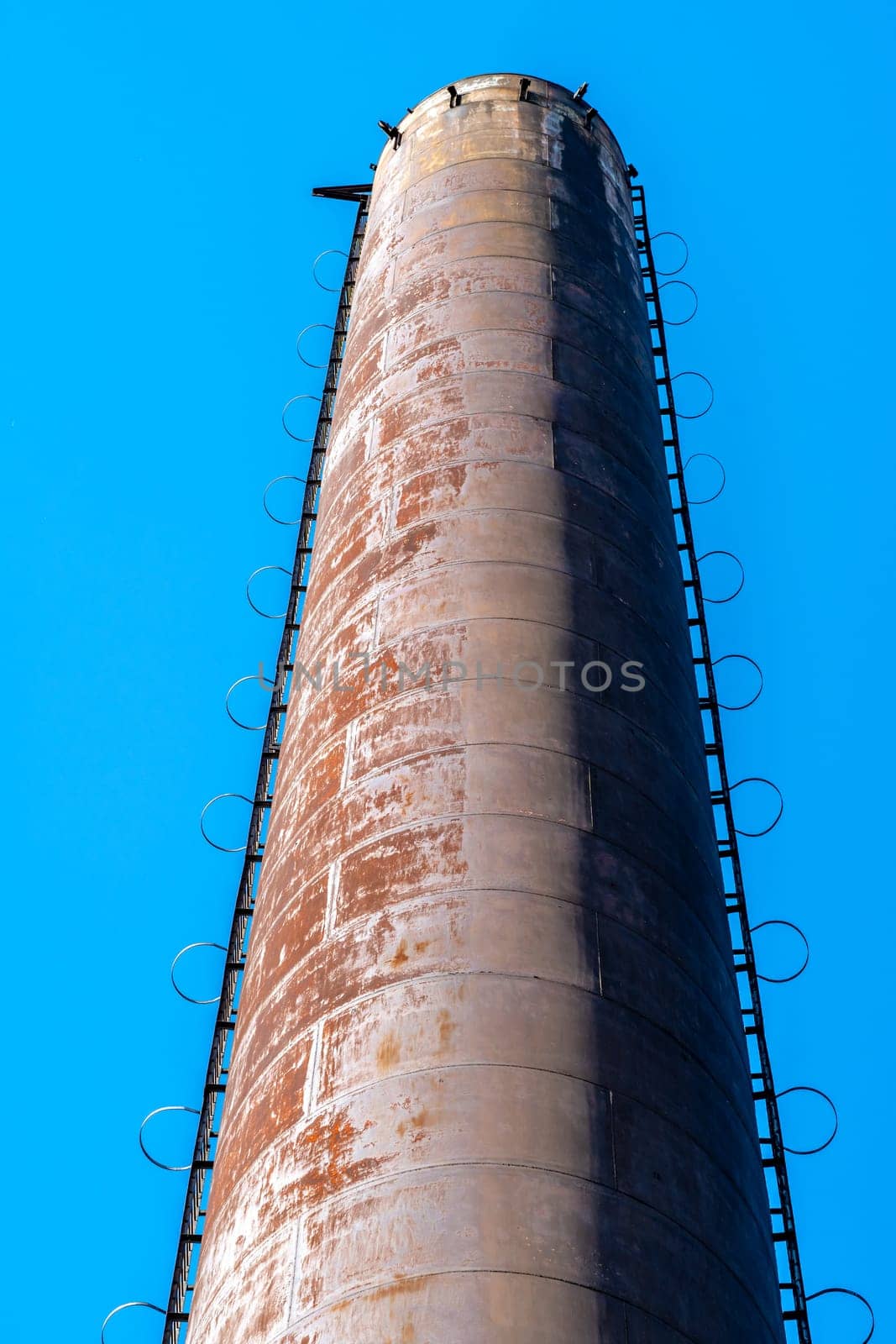 Tall rusty metal chimney with a ladder on the side stands out against a clear blue sky, showcasing industrial architecture