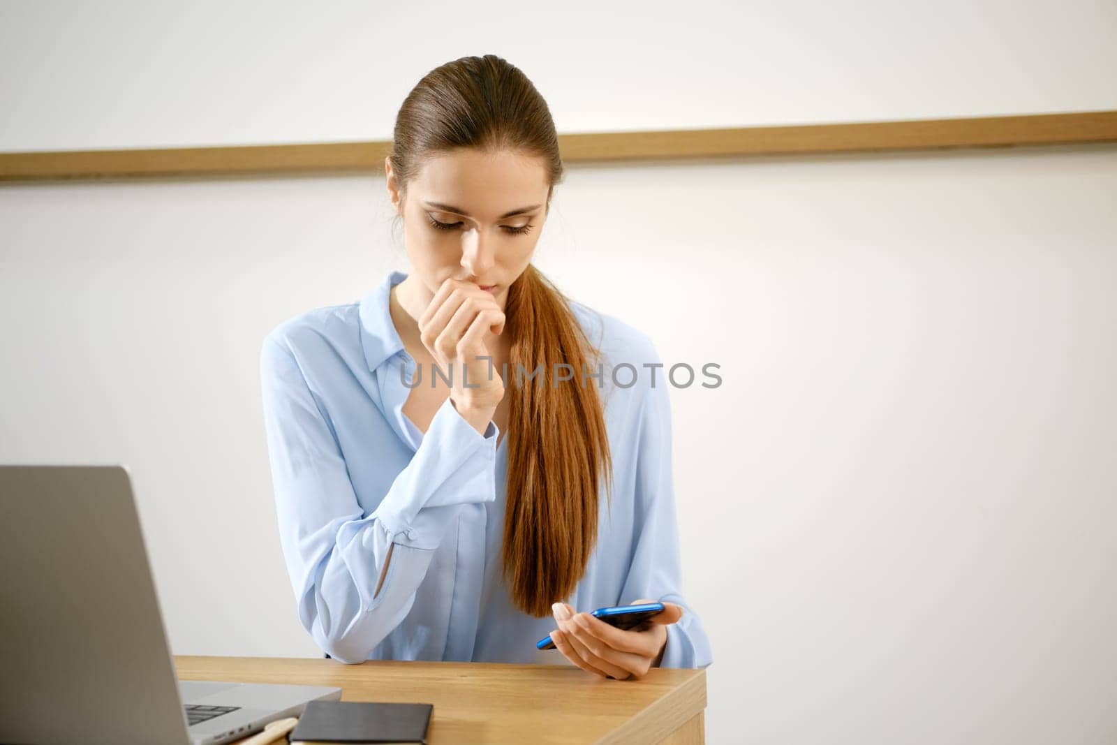 A woman with long brown hair sits at a wooden desk, gazing thoughtfully at her mobile phone.