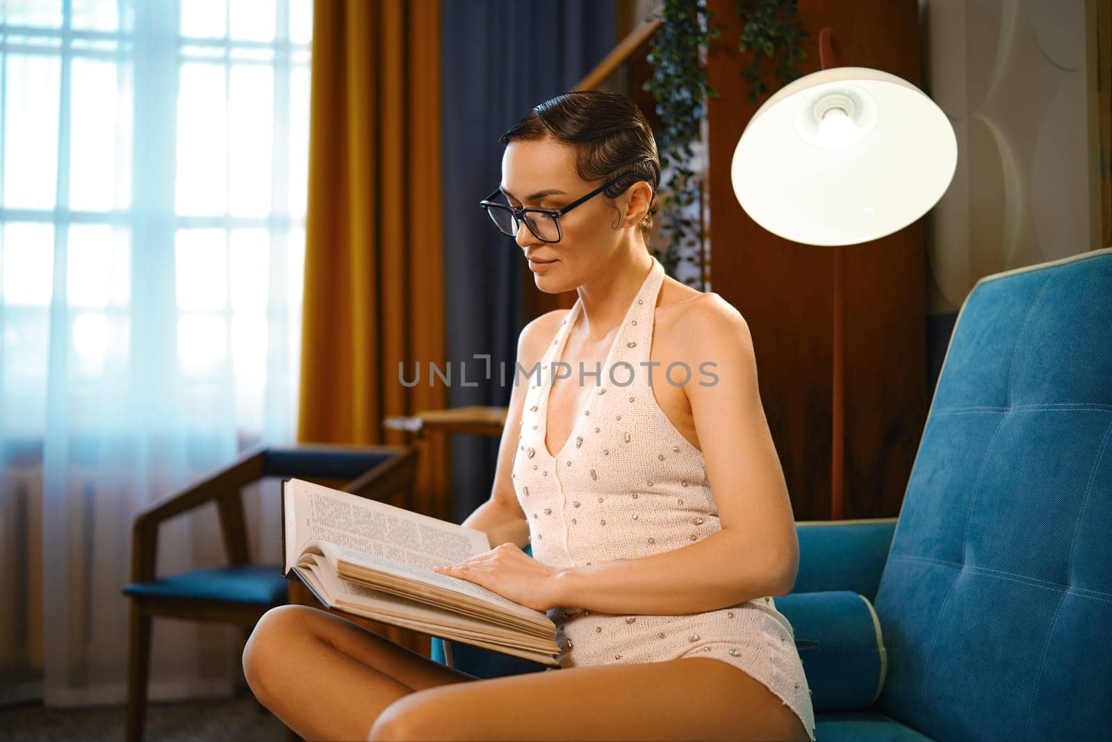 Relaxed woman with old-fashioned hairdo and knitted body suit sits on a sofa and reading book in cozy living room