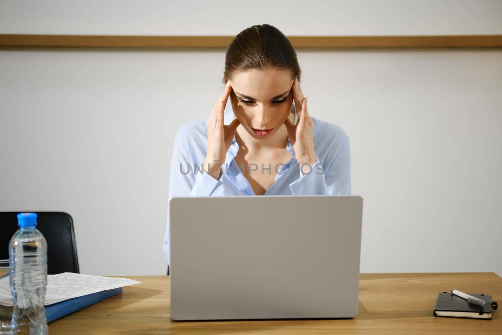 A woman sits at a desk in a contemporary office, visibly stressed as she rubs her temples while looking at a laptop screen.