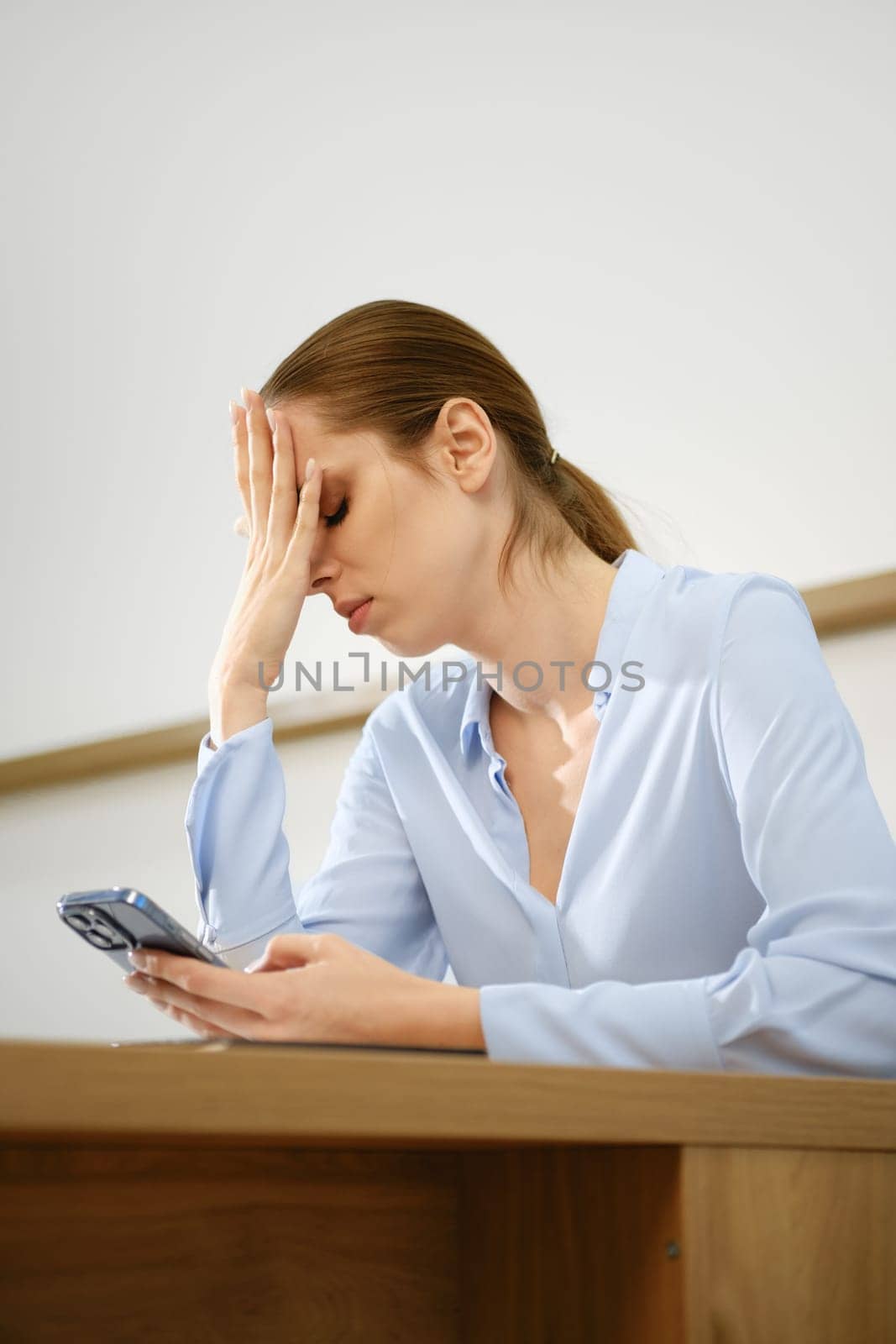 A woman sits at a wooden desk, resting her forehead on her hand while staring at her smartphone with a distressed expression. The bright room highlights her frustration and worry.