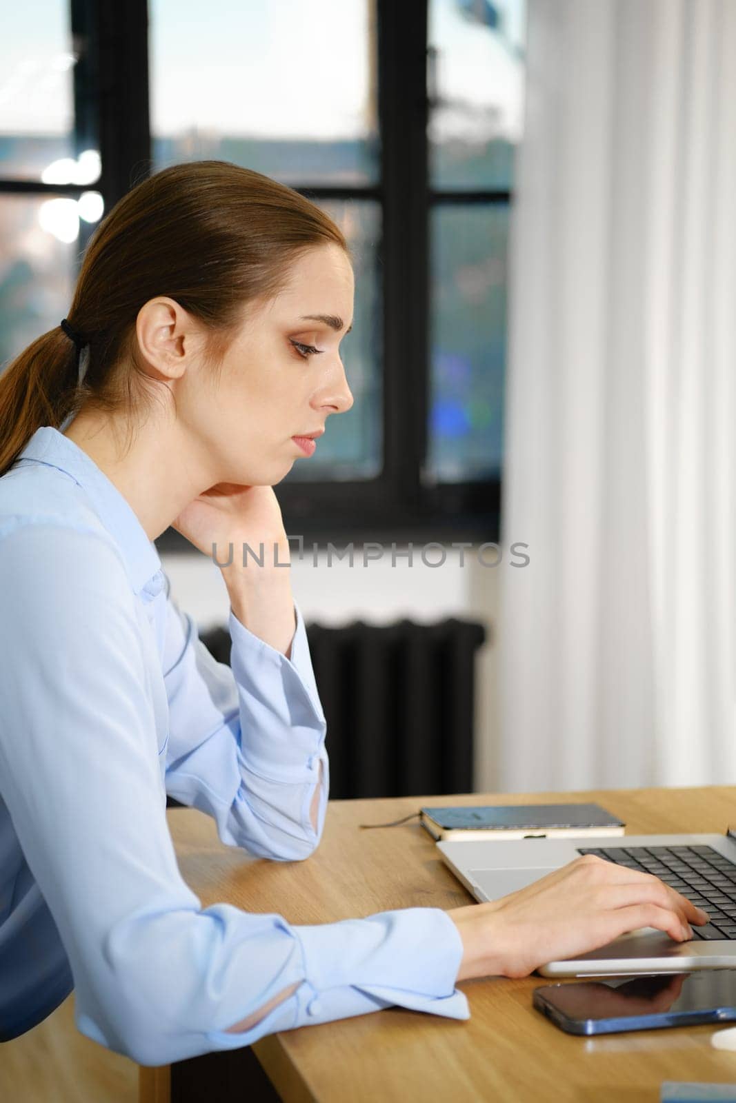 A woman in a light blue blouse is focused on her laptop, seated at a wooden desk in a well-lit office. Natural light streams in through large windows, enhancing the productive atmosphere.