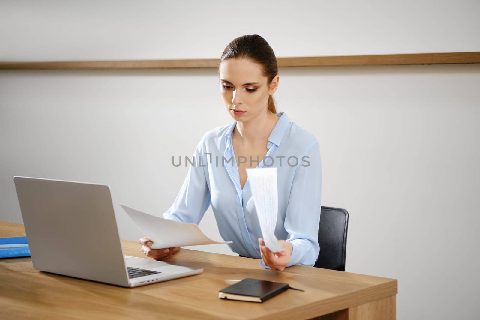 A woman is attentively examining important documents at a wooden desk, with a laptop open in front of her.