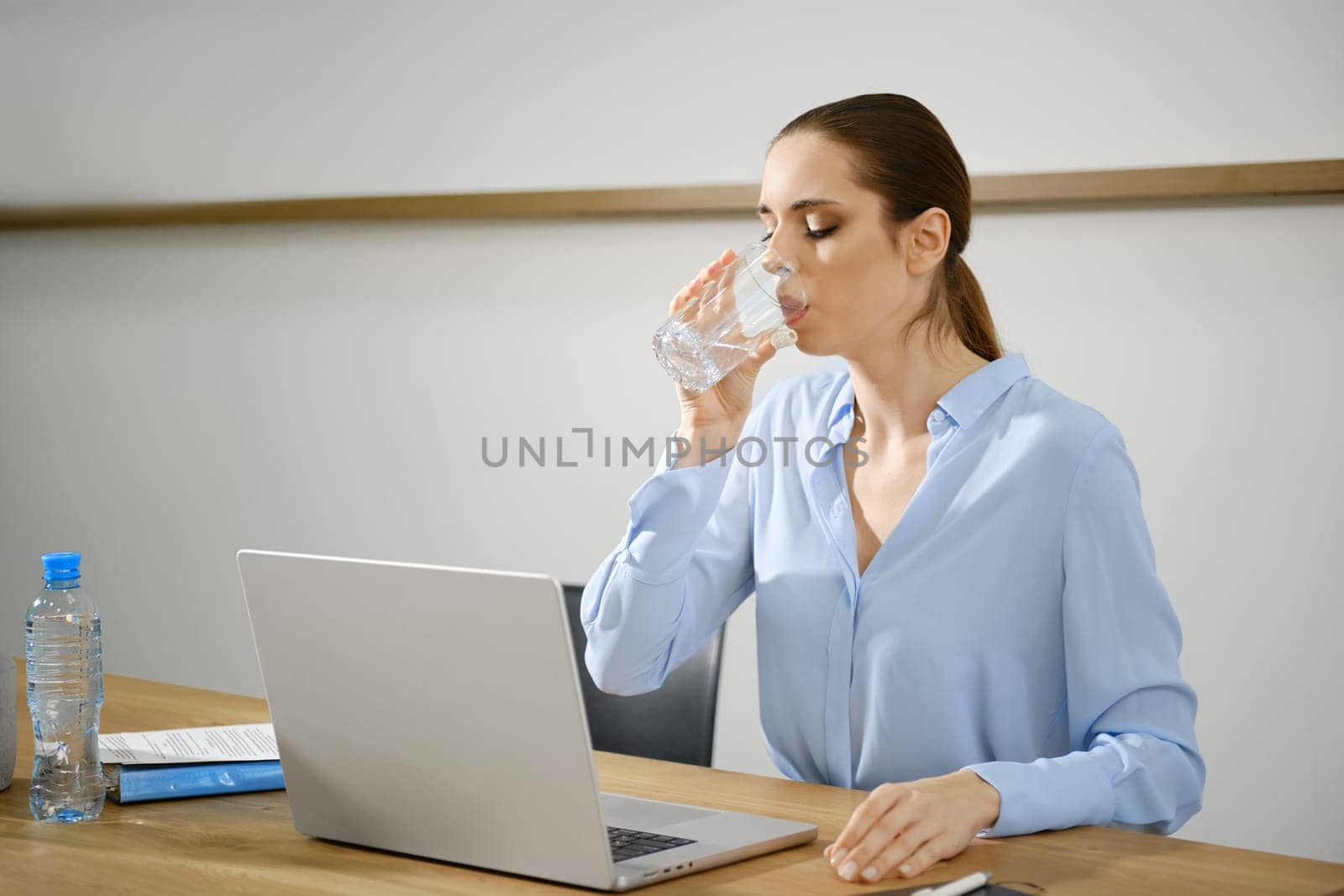 A woman in a light blue blouse pauses to sip water from a glass while working on her laptop at a wooden desk in a modern office environment, maintaining focus and hydration during her tasks