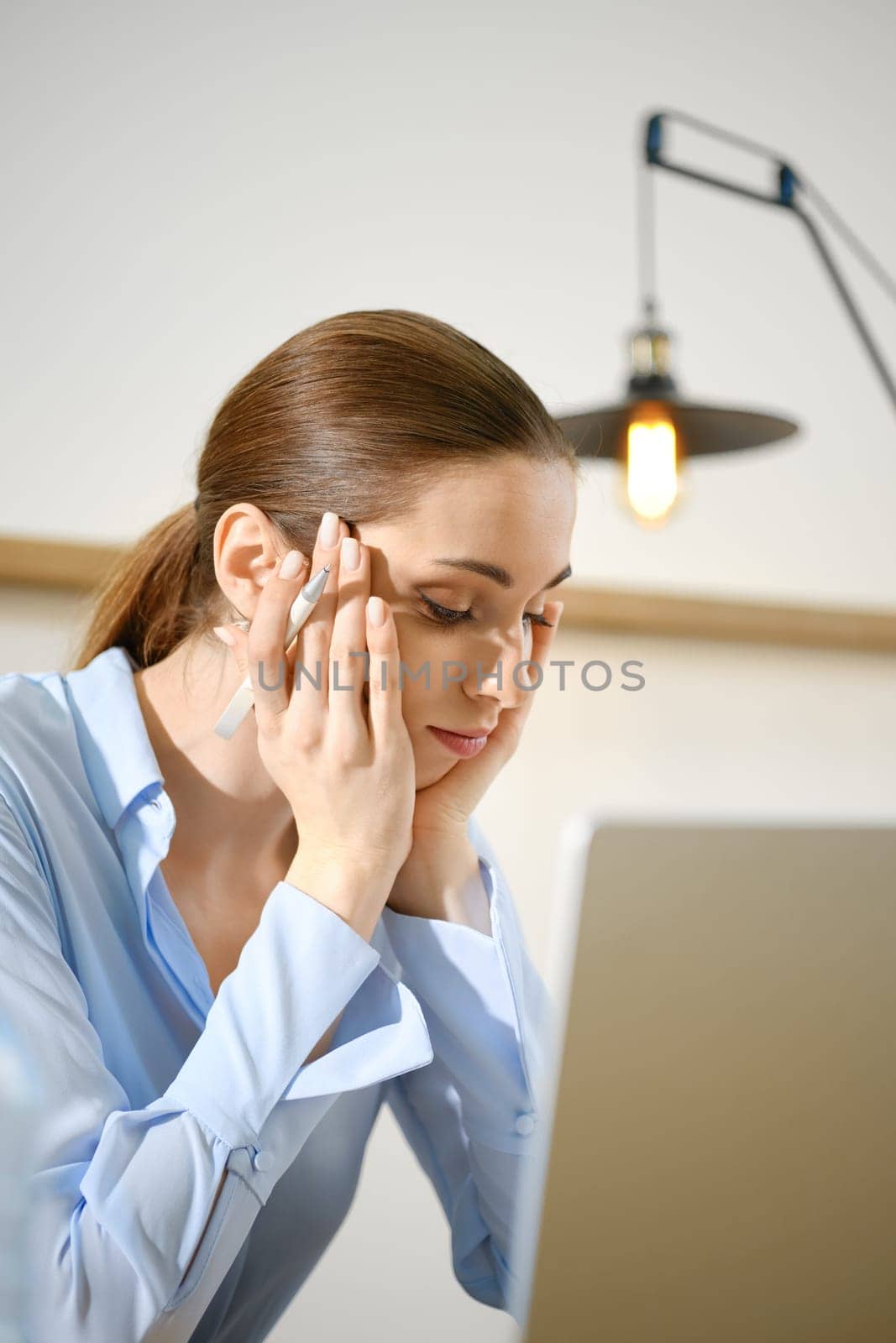 The woman is seated at her desk, visibly stressed as she rests her head on her hands.